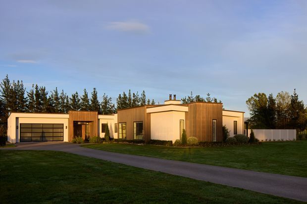 Single-storey contemporary residence viewed from the front, set within a broad landscaped lawn. The home has a low, horizontally composed form with flat roofs, pale rendered walls, warm vertical timber cladding at the central volume, large dark-framed windows, and an integrated double garage. A long paved driveway leads toward the house, with mature trees and fencing behind it.