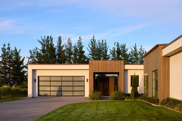 Front exterior view of a contemporary single-storey residence with a low, flat-roofed composition. The facade combines smooth light rendered walls with warm vertical timber cladding around the recessed entry, a large glazed sectional garage door, narrow vertical windows, landscaped lawn, and mature trees beyond.
