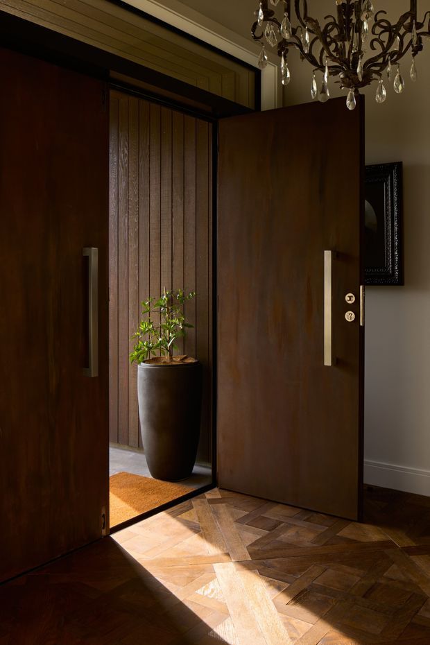 A dramatic, warmly lit entryway viewed from inside, featuring large dark timber double doors with long vertical metal handles, a matching timber-clad vestibule, a tall potted plant, and richly patterned parquet flooring. A crystal chandelier and framed wall artwork add a refined, traditional character.