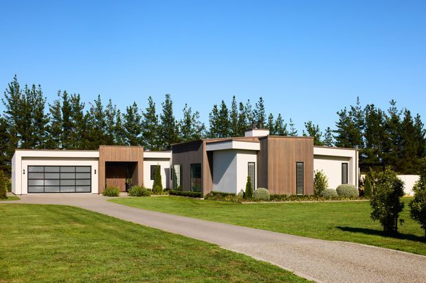 Single-storey contemporary residence viewed from the front, with a low, horizontally composed form, flat rooflines, crisp white rendered walls, dark charcoal feature volumes, vertical timber cladding, extensive glazing, and an integrated double garage. The house sits behind a broad lawn and curved driveway, with mature trees forming the backdrop.