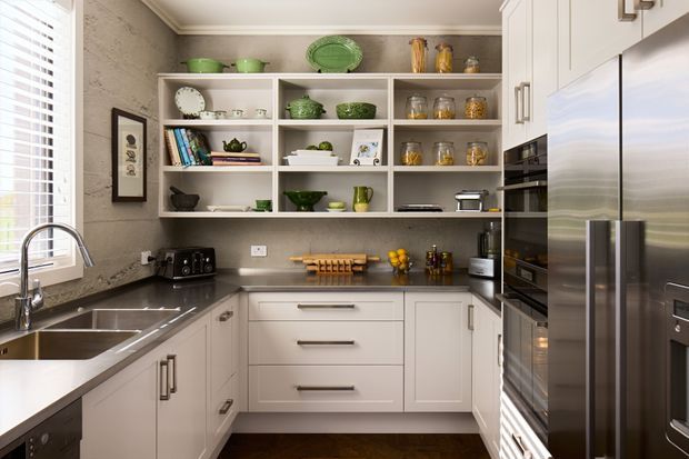 A compact, well-appointed contemporary kitchen with white shaker-style cabinetry, dark stone benchtops, stainless-steel appliances, and open display shelving. The space includes a double-bowl sink beneath a window, integrated oven and microwave, a large refrigerator, and warm decorative ceramics and timber accessories. Natural light enters through white plantation shutters, while a textured grey backsplash or wall finish adds contrast.