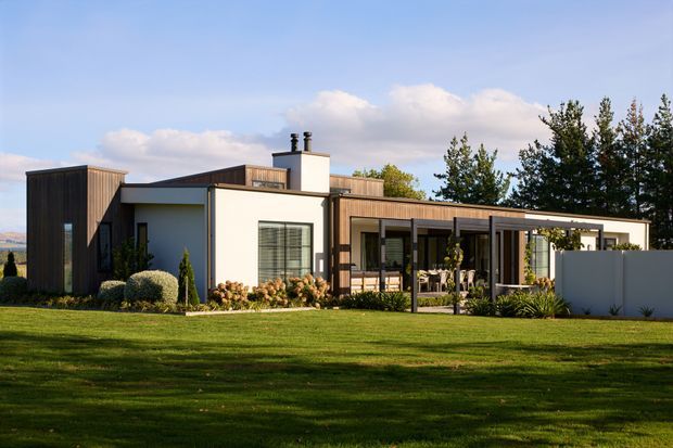 Single-storey contemporary residence viewed from the landscaped front lawn, composed of crisp white rendered volumes, dark vertical timber-clad sections, and a long glazed outdoor living area beneath a flat-roofed pergola. The home has a low, horizontal profile, broad windows and doors, a chimney, and carefully planted garden beds.