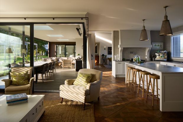 A bright, open-plan interior combining a contemporary kitchen, dining area and living space. The kitchen features a large island with a pale stone-look benchtop, white cabinetry, timber bar stools and two oversized woven pendant lights. Dark herringbone timber flooring runs throughout, while black-framed glazed doors separate the dining area from the living room. Neutral upholstered armchairs, a textured rug and abundant daylight create a warm, inviting atmosphere.