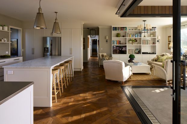 Open-plan residential interior combining a white kitchen with a large island and integrated seating, and an adjacent living area with upholstered armchairs, a sofa, coffee table, and full-height shelving. The space has warm parquet flooring, white paneled cabinetry and walls, pendant lighting, and abundant natural light from large glazed doors or windows. A partial upper-level or stair opening is visible above the living area.