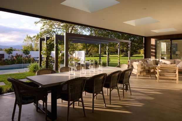 A spacious covered outdoor entertaining area with a long dark dining table and woven chairs, opening onto a landscaped garden and swimming pool. A relaxed lounge setting sits beneath a broad flat roof with recessed skylights, while dark-framed glazed openings and a pergola-like structure connect the terrace to the garden. Warm afternoon light creates long shadows across the concrete floor.