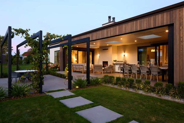 Single-storey contemporary home viewed from the landscaped garden, with a broad covered outdoor entertaining area opening directly to the interior. The facade combines dark timber cladding with large black-framed glazed openings, recessed ceiling lighting, an outdoor dining setting, lounge seating, and a visible kitchen or bar area. A paved stepping-stone path crosses the lawn, while a pergola-like black structural frame supports climbing planting along the garden edge.