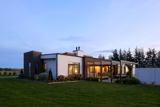 A single-storey contemporary home photographed at dusk, set within a broad landscaped lawn. The building combines white rendered wall volumes with dark vertical timber-clad sections, a low flat roofline, large glazed openings, and a covered outdoor living area supported by slender posts. Warm interior lighting is visible through the windows, with a white boundary wall and mature trees behind the property.