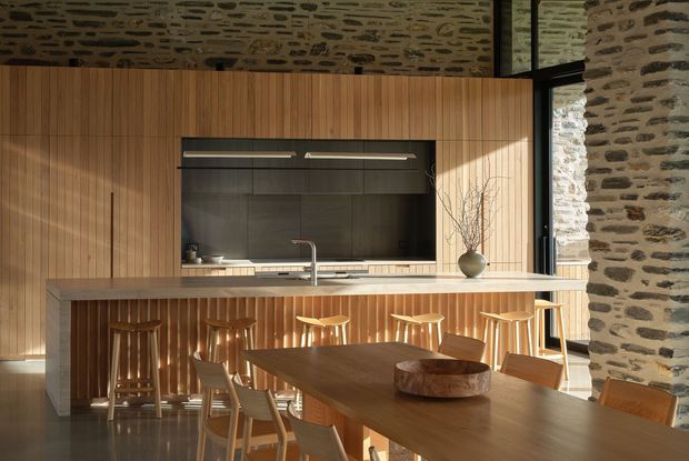 Warm contemporary open-plan kitchen and dining area set within an existing rough stone masonry shell. Vertical timber wall panelling wraps the kitchen cabinetry and island, with a dark recessed cooking zone, pale stone benchtop, timber stools, and a large timber dining table. Exposed stone walls and large black-framed glazing create a strong contrast between rustic structural fabric and refined modern joinery.