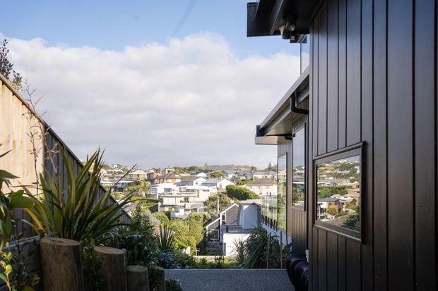 Exterior view along a narrow landscaped walkway beside a contemporary dark-clad house, looking downhill across a suburban neighbourhood. The building has vertical dark cladding, simple projecting eaves, and reflective glazing, with coastal-style planting and timber edging in the foreground.