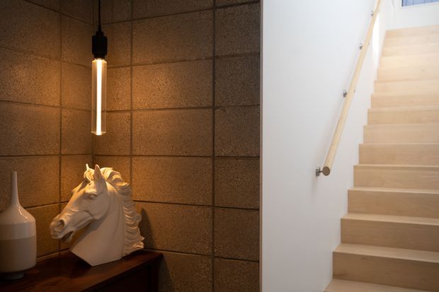 Interior view of a stairway beside a warmly lit feature wall. Pale timber stairs with a simple timber handrail rise along a white wall, while the adjacent wall is clad in dark textured square panels and features a slim pendant light above a decorative white horse sculpture on a dark console.