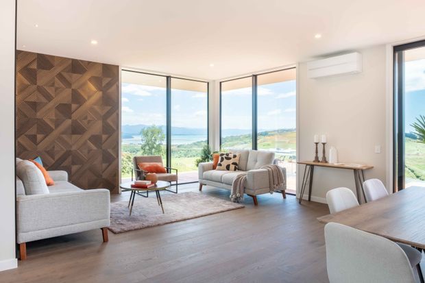 Bright contemporary open-plan living and dining area with expansive corner glazing overlooking a green landscape. The room features pale upholstered seating, a timber coffee table, a large rug, a dark geometric timber feature wall, and a timber dining table with upholstered chairs. Recessed ceiling lighting and a wall-mounted split-system air conditioner complete the clean, refined interior.
