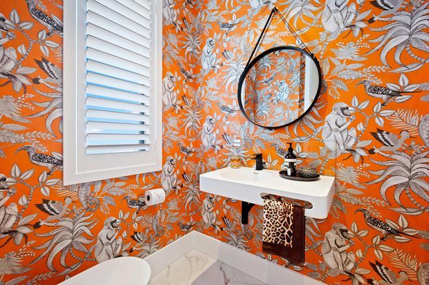 A compact, highly decorative bathroom featuring vivid orange botanical and animal-print wallpaper, white plantation shutters, a wall-mounted white basin with black tapware, a round black-framed mirror, and pale marble-look floor and wall surfaces. A small leopard-print stool adds a bold accent beneath the vanity.