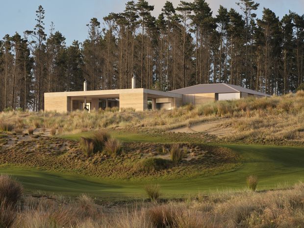 A low-profile contemporary single-storey residence set into a gently sloping, sandy grass-covered site at the edge of a tall pine forest. The home has pale timber-toned horizontal cladding, broad dark-framed glazing, a projecting flat-roofed living wing, and a low hipped or shallow pitched roof over the adjoining volume. Native grasses and landscaped mounds surround the building.