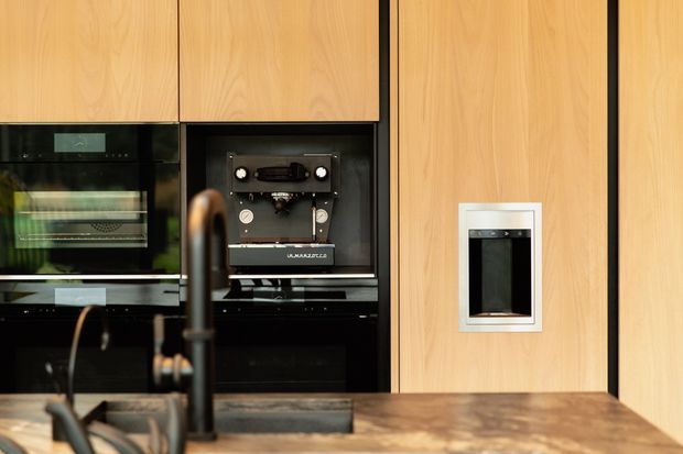 Close-up view of a contemporary kitchen with full-height light timber cabinetry, integrated black appliances, a built-in espresso machine, and a dark stone island benchtop. A matte black gooseneck tap and sink are visible in the foreground, with strong tonal contrast between the warm wood and black appliance wall.