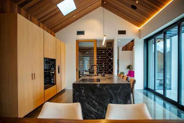 A refined open-plan kitchen and dining area with a dramatic dark stone island, integrated light-wood cabinetry, extensive wine storage, and floor-to-ceiling glazing. The room features a warm timber-lined vaulted ceiling with a skylight, linear pendant lighting, and polished flooring.