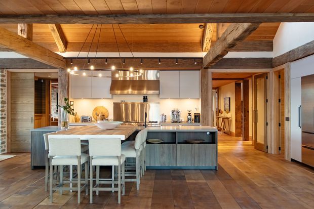 A spacious rustic kitchen with a large central island, integrated dining seating, stainless-steel appliances, pale cabinetry, and warm timber surfaces. Exposed heavy timber beams and posts frame the open-plan space, with a timber ceiling, broad timber flooring, and hallways leading to adjoining rooms. Contemporary linear pendant lighting contrasts with the traditional structural fabric.