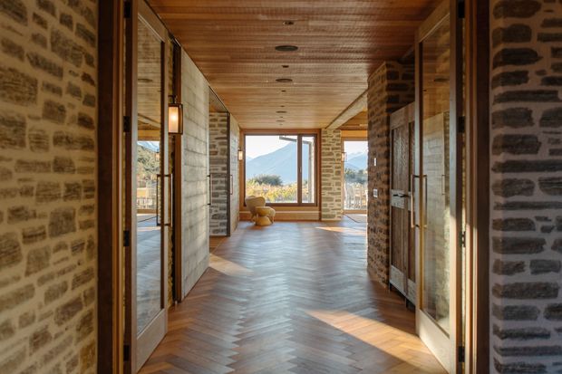A warm, elongated interior passage or entry hall with exposed rough stone walls, extensive timber wall and ceiling lining, and a herringbone timber floor. Glazed and timber-framed doors or partitions line both sides, while a large window at the far end provides views toward a mountainous landscape and brings in strong natural light. The space has a rustic, lodge-like character with carefully detailed built-in glazing and masonry.