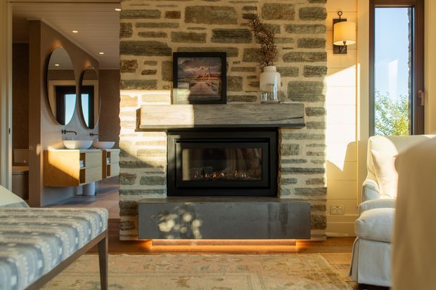 Warm, refined living room centred on a substantial natural stone fireplace with a black built-in firebox and illuminated hearth base. The room includes a pale upholstered sofa, patterned rug, timber flooring, a patterned upholstered bench, and a timber vanity or console visible to the left beneath two round mirrors. Strong afternoon sunlight enters through a tall window, highlighting the textured stonework and soft neutral finishes.