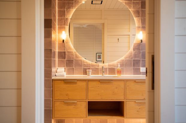A warm, compact bathroom vanity viewed through a doorway. The room features a timber floating vanity with drawers and an open central shelf, a light benchtop, a large round mirror set within a square field of earthy brown tiles, warm wall sconces, and pale horizontal wall panelling.