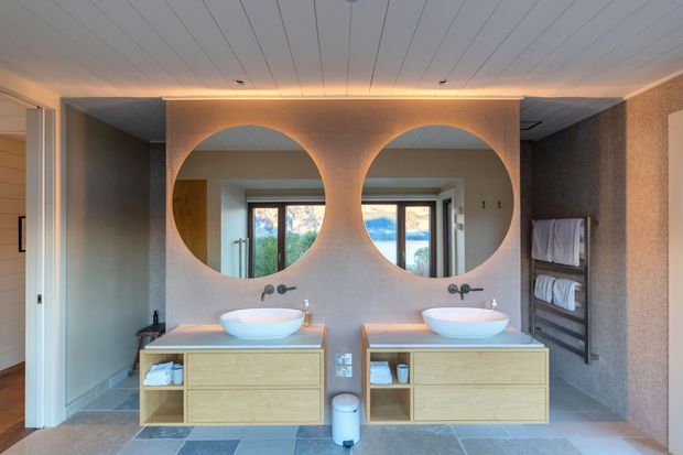 A warm contemporary bathroom vanity area with two floating timber vanities, white vessel basins, dark wall-mounted taps, and large circular mirrors set into a sculptural curved feature wall. A glass shower enclosure and folded towel shelving are visible, with natural light entering through the rear glazing. The room has a pale tiled floor, textured neutral walls, and a linear timber-look ceiling with concealed warm lighting.