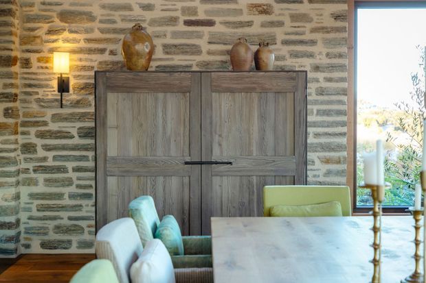 Warmly styled dining area featuring a large pale stone dining table with upholstered seating, a substantial dark timber cabinet, and a natural irregular-stone feature wall. A tall window provides views to greenery, while wall-mounted and table lighting add a cozy ambience. Decorative ceramic vessels sit on the cabinet.