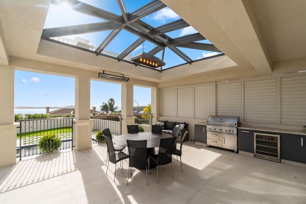 Bright enclosed outdoor entertaining area with a retractable glazed roof, tiled flooring, dining setting, built-in outdoor kitchen, barbecue, beverage fridge and louvred shutters. The open side overlooks a pool and coastal waterfront, creating an airy indoor-outdoor living space.