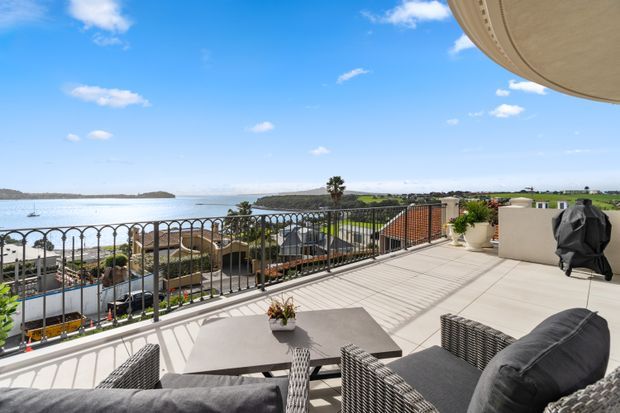A spacious elevated outdoor terrace overlooking a bay or waterfront, furnished with dark woven lounge seating and a dining table. The terrace has pale tiled paving, ornate metal balustrades, potted plants, and a curved overhanging roof or soffit, with a bright open coastal landscape beyond.