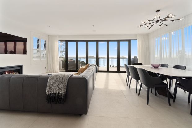 Bright, open-plan contemporary living and dining area with a large dark sectional sofa, linear fireplace, white walls, pale tiled flooring, full-height glazed doors and windows, and a black sculptural chandelier. The dining zone overlooks a waterfront or pool-like setting beyond the railing.