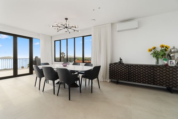 Bright, contemporary dining room with a large rectangular dining table and dark upholstered chairs, set against expansive black-framed glazing opening to a balcony or terrace. A sculptural branching pendant light hangs above, while full-height curtains, a patterned dark sideboard, pale flooring, and a wall-mounted split-system air conditioner complete the space. A coastal or elevated view is visible beyond the glazing.