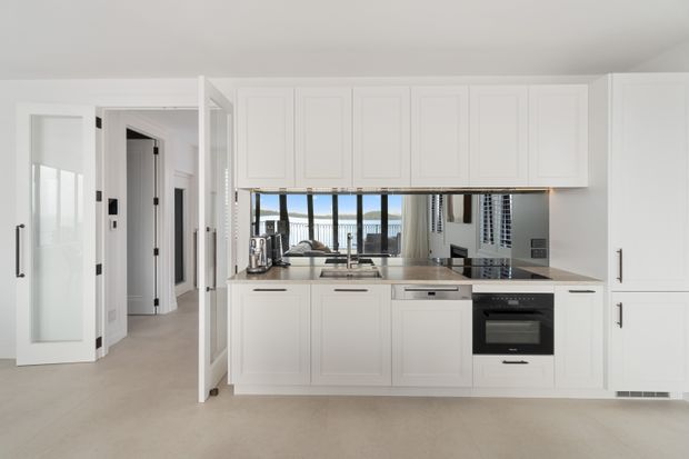 Bright, compact white kitchen with extensive Shaker-style cabinetry, a dark stone-look benchtop, stainless-steel sink and appliances, and a reflective splashback opening toward a living area with large windows. A hallway and glazed interior door are visible to the left.
