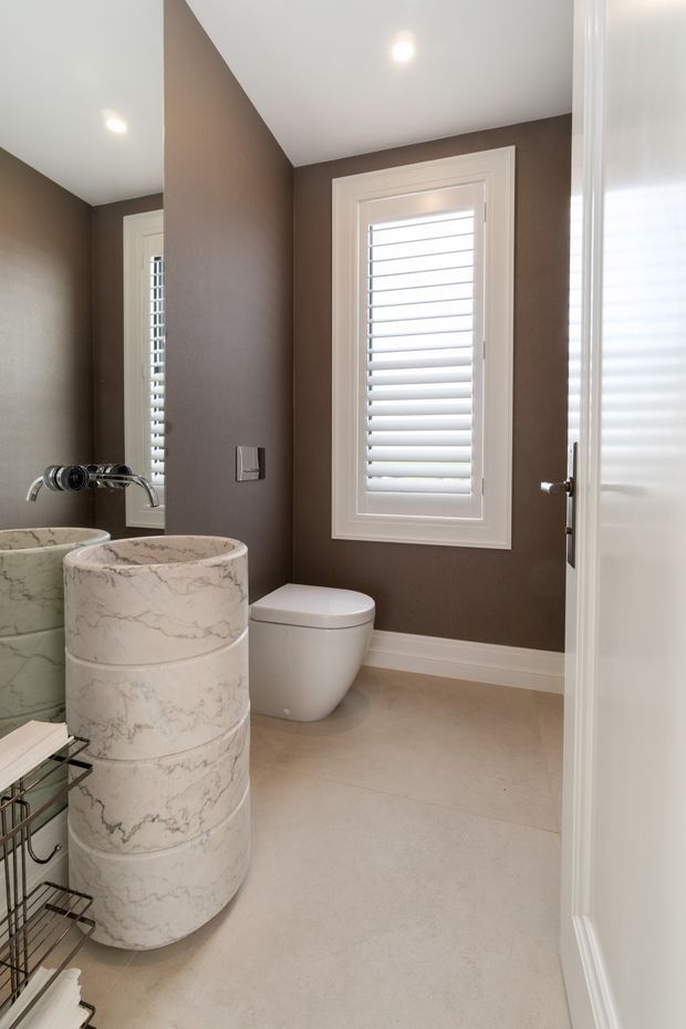 A compact, refined bathroom viewed from the doorway, featuring a sculptural cylindrical marble-patterned basin, wall-mounted chrome tapware, a white toilet, pale tiled flooring, taupe-brown walls, white trim, and plantation-shuttered windows. A large mirror reflects the basin and fittings.