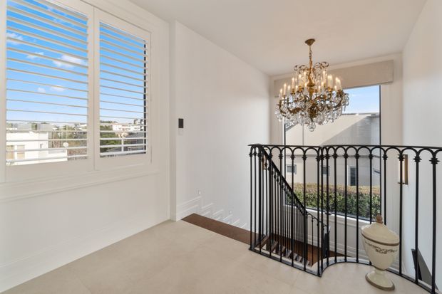 Bright upper-level landing or hallway overlooking a staircase, featuring white walls, plantation shutters, beige carpet, dark timber stair trim, ornate black metal balustrades, and a prominent traditional crystal chandelier. Large windows provide views of neighbouring rooftops and greenery.