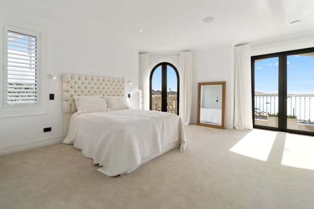 Bright, spacious bedroom with a cream upholstered button-tufted bedhead, white bedding, pale carpet, white walls and curtains, and black-framed glazed doors opening to a balcony. A tall arched window and plantation shutters add architectural character, while strong sunlight creates a warm, airy atmosphere.