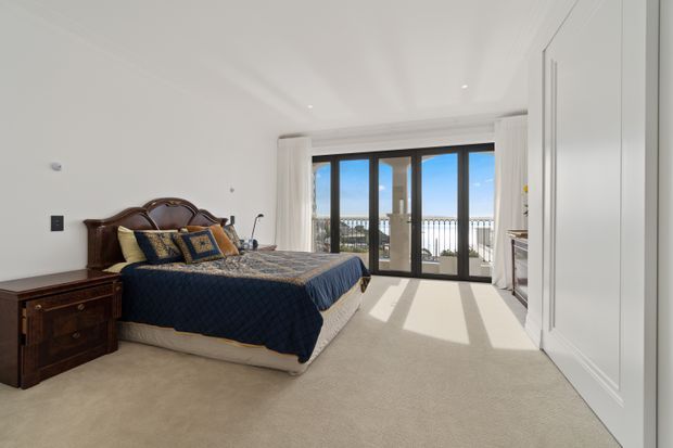 Bright, spacious bedroom with a traditional dark wood bed and bedside table, navy and patterned bedding, soft beige carpet, white walls and decorative wall panelling. Full-height glazed doors open to a balcony, bringing in strong natural light and coastal or elevated views.