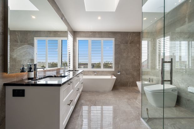 Bright, spacious contemporary bathroom with a freestanding white bathtub, floating white vanity with a dark stone benchtop, large-format grey stone-look wall and floor finishes, frameless glass shower enclosure, and a separate toilet. Plantation shutters and a large skylight provide abundant natural light.