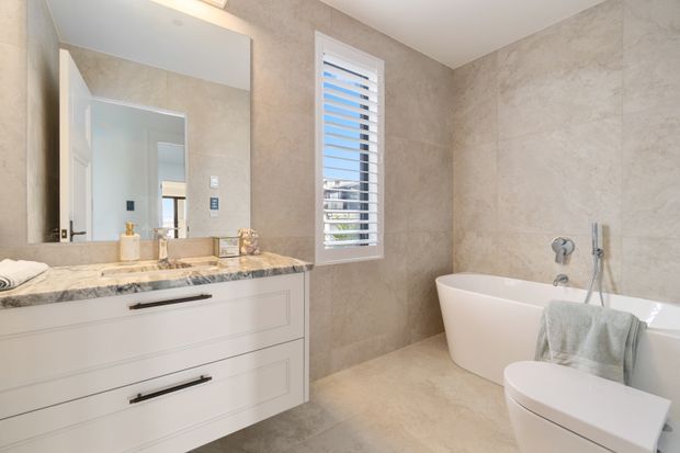 Bright contemporary bathroom featuring a freestanding white bathtub, wall-mounted white vanity with dark handles, marble-look benchtop, large frameless mirror, beige stone-look wall and floor tiling, and a white shuttered window.