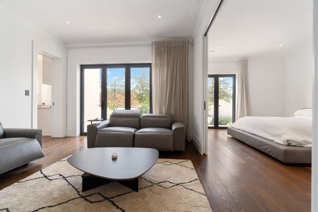 Bright contemporary open-plan living room and bedroom viewed together, featuring white walls and decorative ceiling cornices, warm timber flooring, full-height black-framed glazing with neutral curtains, a grey leather sofa, low rounded black coffee table, and a cream geometric rug. A bedroom with a low upholstered bed is visible through a large internal opening or glazed partition.