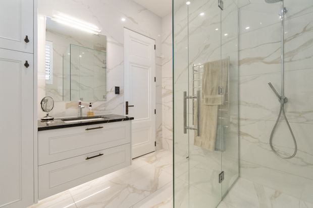 Bright, refined bathroom featuring a white vanity with dark stone benchtop, large marble-look wall and floor tiles, a frameless glass walk-in shower, polished chrome fixtures, and recessed lighting. A mirrored vanity wall and white paneled door reinforce the clean, upscale aesthetic.