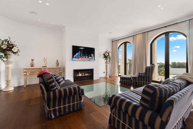 Elegant formal living room with dark timber flooring, white walls and ceiling, a marble-look fireplace surround, and a wall-mounted television. Multiple navy and cream striped upholstered armchairs and sofas are arranged around a glass coffee table. Tall arched black-framed glazed doors and windows open to a bright exterior view, with full-height beige curtains and traditional decorative console furniture adding a refined classic character.