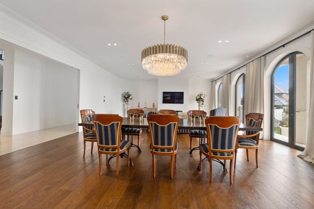Elegant formal dining room with a long dark timber dining table surrounded by upholstered, timber-framed dining chairs. The room has polished timber flooring, white walls and ceiling with decorative cornices, tall arched glazed doors, full-height curtains, recessed lighting, and a large ornate crystal chandelier.