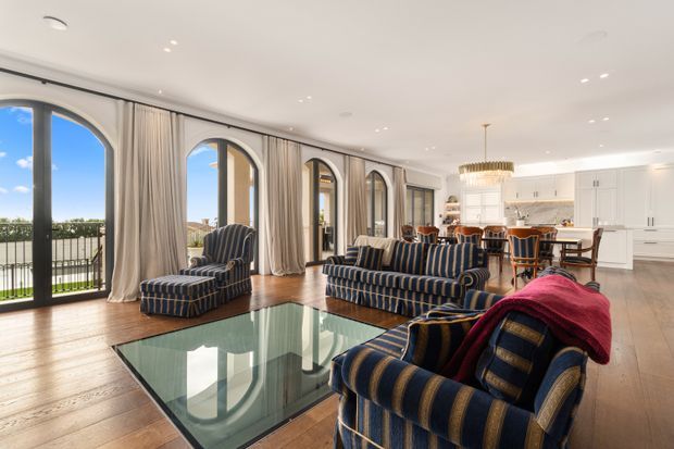 Bright open-plan living room and kitchen with a polished traditional interior. The room features large arched floor-to-ceiling glazed doors with full-height curtains, warm timber flooring, striped navy upholstered sofas and armchairs, a large glass coffee table, and a white kitchen with a central island and pendant chandelier. A balcony and exterior view are visible beyond the glazing.