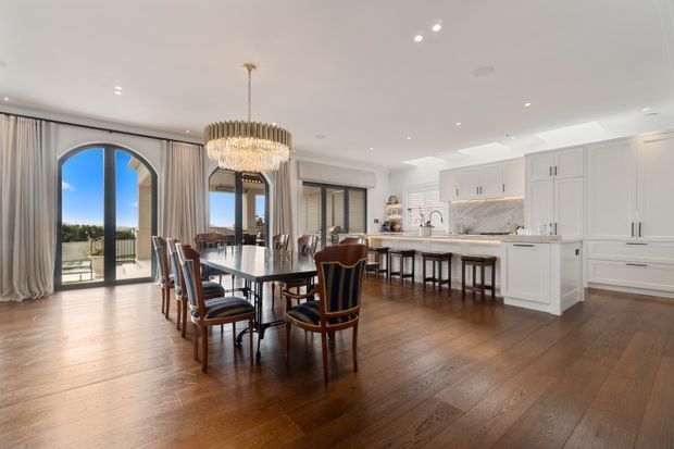 A bright, spacious open-plan dining and kitchen interior with a large dark timber dining table, upholstered dining chairs, a crystal chandelier, white classical-style cabinetry, marble-look benchtops and splashback, and warm timber flooring. Tall arched glazed doors open toward an exterior terrace, while full-height curtains and recessed ceiling lighting complete the refined setting.