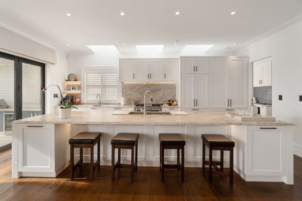 Bright, spacious white kitchen with a large central island and seating for four. The room features shaker-style cabinetry, a pale stone benchtop, a marble-look splashback, dark timber flooring, skylights, recessed lighting, plantation shutters, and black-framed glazed doors.