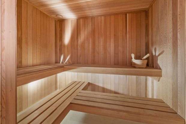 A compact traditional sauna lined throughout with warm timber vertical wall panelling and a timber slatted ceiling. Tiered wooden bench seating wraps around the room, with subtle concealed lighting and a small sauna bucket and ladle on the upper bench. The space is warmly lit and uncluttered.