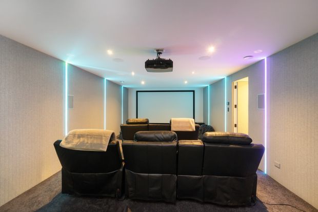 A dedicated home cinema room viewed from behind a row of black reclining theatre seats. The room features a front projection screen, ceiling-mounted projector, dark carpet, textured grey walls, and cyan and purple LED accent lighting around the perimeter. A doorway is visible on the right.