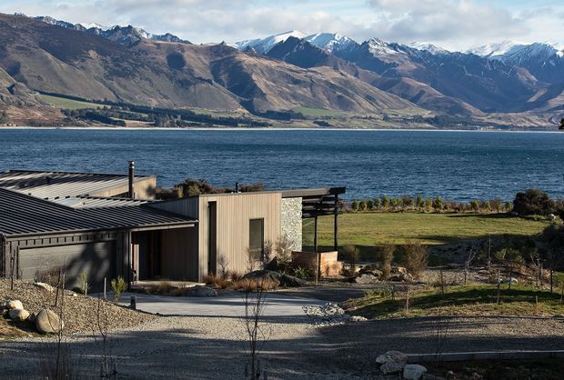 Exterior view of a low-slung contemporary residence beside a large lake, with a corrugated metal roof, timber-toned cladding, stone feature wall, broad lawn and mountain ranges beyond. The building sits in a spacious, sparsely landscaped setting with a gravel driveway and outdoor terrace structure.