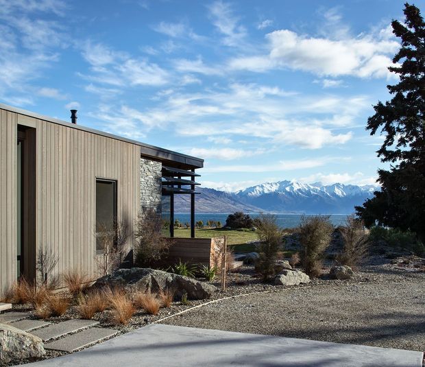 Exterior view of a contemporary single-storey building clad in vertical timber boards, with a stone wall section and deep roof overhang. The landscaped foreground uses gravel, large rocks, drought-tolerant grasses and shrubs, while the property overlooks a lake with snow-capped mountains in the distance.
