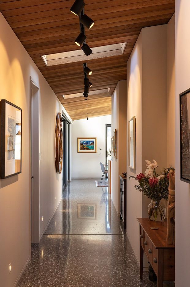 A long, refined residential hallway with polished terrazzo flooring, warm timber-lined ceiling, recessed skylights, and a row of black directional track lights. White walls are lined with framed artwork, doors, and built-in or console storage. A timber console with flowers occupies the right foreground, while the corridor opens toward a bright seating or living area in the distance.