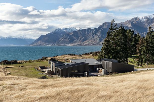 A low-profile contemporary residence set in a rugged lakeside landscape. The dark, rectilinear building appears to use corrugated metal cladding and flat or very low-pitched roof forms, with large windows and several connected wings. Dry golden grass, evergreen trees, turquoise water, and dramatic snow-capped mountains dominate the surrounding setting.