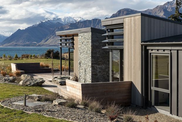 Exterior view of a contemporary lakeside mountain residence, featuring dark vertical cladding, pale timber-look wall panels, stacked natural-stone walls, extensive glazing, and flat roof planes. The landscaped foreground includes stone-edged garden beds, gravel, timber planter walls, and a paved outdoor sitting area. A lake and dramatic snow-capped mountains form the distant setting.